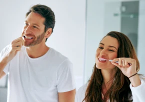 Um casal escova os dentes lado a lado em frente ao espelho, sorrindo e demonstrando cuidado com a saúde bucal.