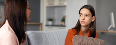 Adolescente sentada no sofá, abraçando um travesseiro, conversa com uma mulher adulta em um ambiente doméstico, representando um momento de apoio e diálogo sobre gravidez na adolescência.